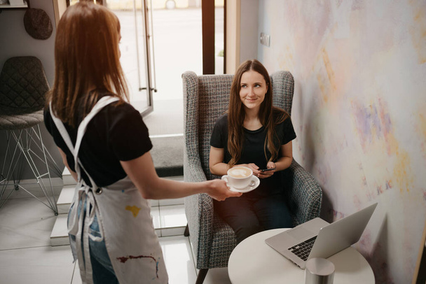 Μια μπαρίστα δίνει έναν λάτε σε μια κοπέλα σε μια καφετέρια. Μια γυναίκα με μακριά μαλλιά που δουλεύει εξ αποστάσεως σε λάπτοπ κρατάει την κοινωνική απόσταση αρπάζει ένα φλιτζάνι καφέ σε ένα καφέ..  - Φωτογραφία, εικόνα