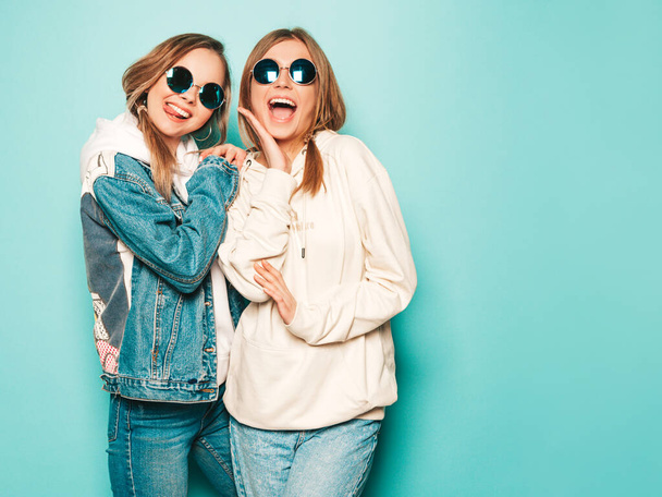 Two young beautiful brunette smiling hipster girls in trendy summer hoodie and jeans jacket clothes. Sexy carefree women posing near blue wall. Trendy and positive models having fun in sunglasses - Foto, imagen