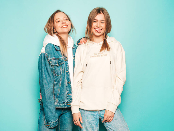 Two young beautiful brunette smiling hipster girls in trendy summer hoodie and jeans jacket clothes. Sexy carefree women posing near blue wall. Trendy and positive models having fun - Photo, Image