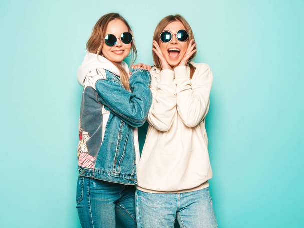 Two young beautiful brunette smiling hipster girls in trendy summer hoodie and jeans jacket clothes. Sexy carefree women posing near blue wall. Trendy and positive models having fun in sunglasses - Fotografie, Obrázek
