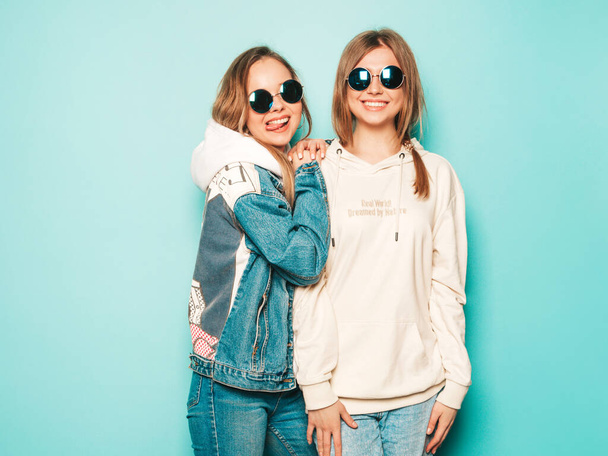 Two young beautiful brunette smiling hipster girls in trendy summer hoodie and jeans jacket clothes. Sexy carefree women posing near blue wall. Trendy and positive models having fun in sunglasses - Fotografie, Obrázek