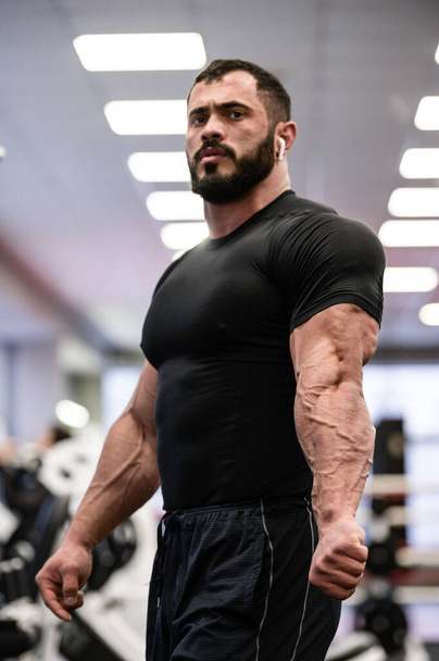 strong bearded sport young man in black sportswear showing his muscle and vein separation on triceps after workout in gym - Photo, image
