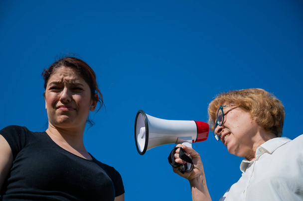 Der Generationenkonflikt. Eine emotionale ältere Frau schreit ihre Tochter per Megafon an. Eine ältere Mutter beschimpft eine Frau mittleren Alters auf einem Lautsprecher auf blauem Hintergrund. - Foto, Bild