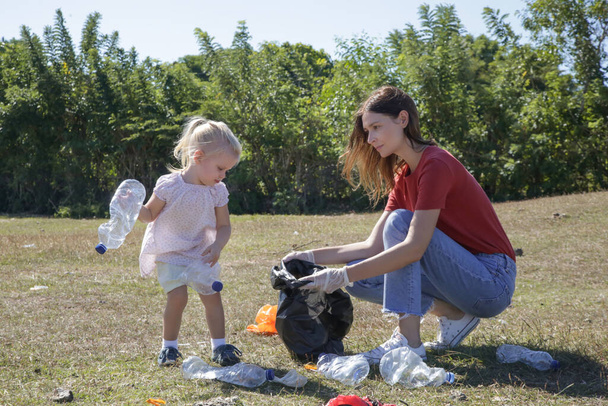 Nuori nainen hakee muovijätettä niityltä pikkutyttönsä kanssa. Roskien kerääminen on kasvatuksellista toimintaa lasten saamiseksi mukaan ympäristönsuojeluun
. - Valokuva, kuva