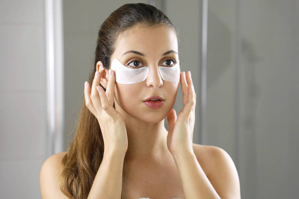 Beauty woman applying under-eye mask looking herself in the mirror in the bathroom. Skin care girl touch patches of fabric mask under eyes to reduce eye bags. - Фото, изображение
