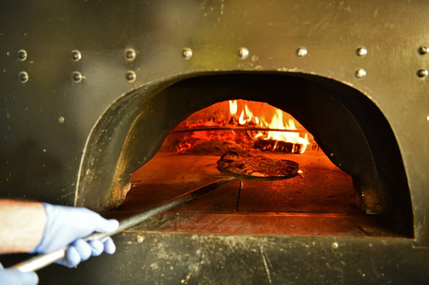 Skilled chef preparing traditional italian pizza  in interior of modern restaurant kitchen with special wood-fired oven. Wearing protective medical face mask and gloves in coronavirus new normal concept - Foto, imagen