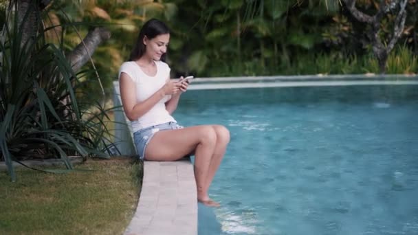 Young woman sits on edge of pool and uses phone for work, greneery on background - Video