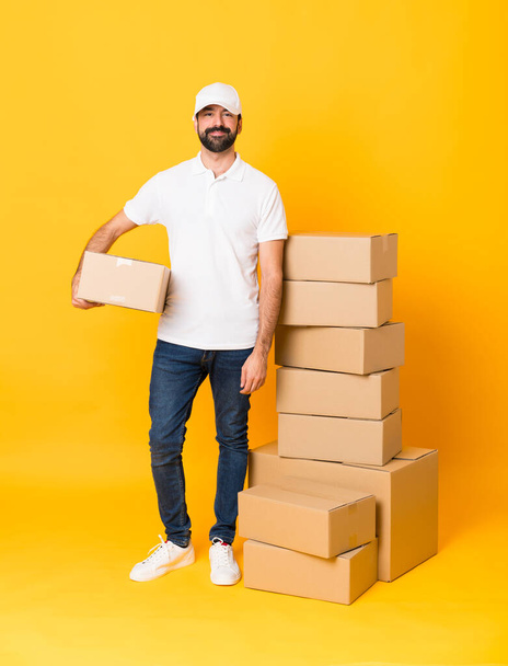 Full-length shot of delivery man among boxes over isolated yellow background keeping the arms crossed in frontal position - Фото, изображение