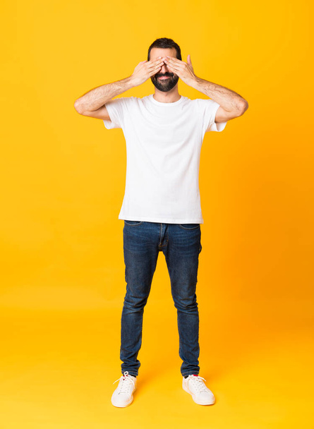 Full-length shot of man with beard over isolated yellow background covering eyes by hands - Photo, Image