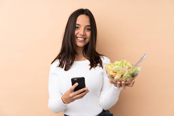 Junges brünettes Mädchen hält einen Salat über isoliertem Hintergrund und sendet eine Nachricht mit dem Handy - Foto, Bild