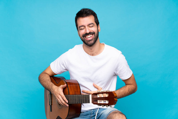 Fiatal férfi gitár felett elszigetelt kék háttér mosolyog sokat - Fotó, kép