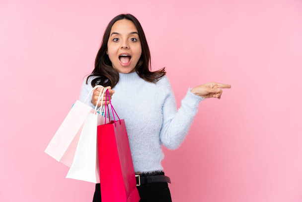 Junge Frau mit Einkaufstasche vor isoliertem rosa Hintergrund überrascht und zeigt mit dem Finger zur Seite - Foto, Bild