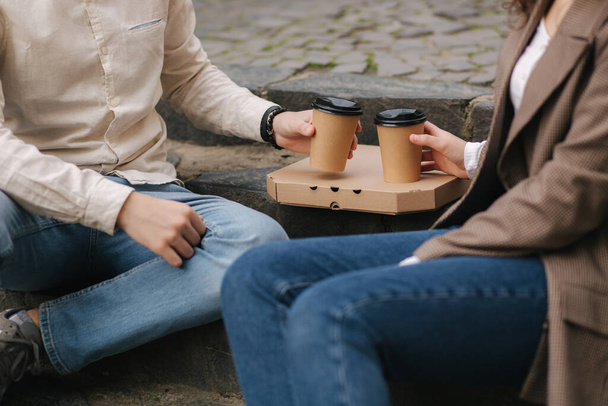 Smiling love couple sitting on the stairs outdoors and hold cups of coffe. Couple has takeaway vegan pizza - Foto, Bild