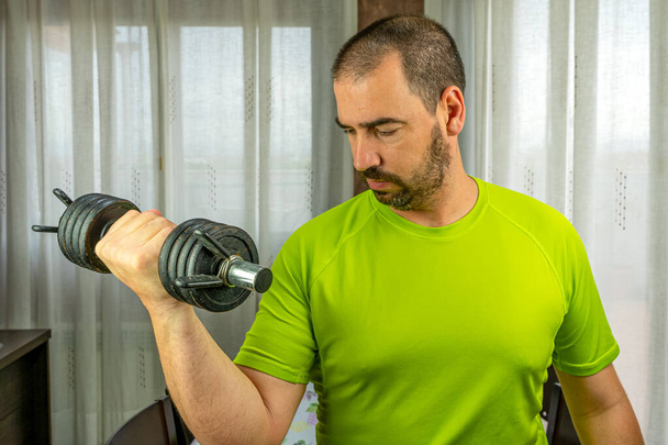 Homme barbu aux cheveux courts en chemise verte faisant des poids dans le salon à la maison. Concept de santé
 - Photo, image