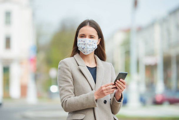 Девушка с длинными волосами в медицинской маске для лица, чтобы избежать распространения коронавируса, использует смартфон на улице. Женщина в маске против COVID-19 носит пальто, смотрит в центр города
. - Фото, изображение