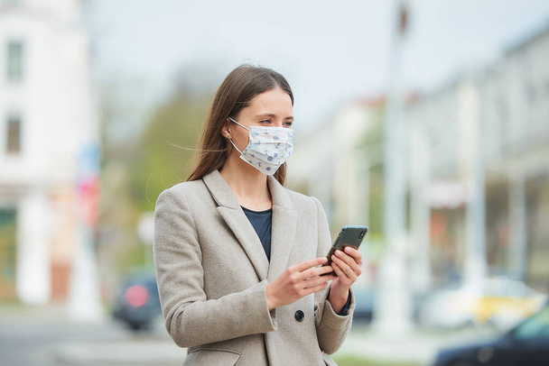 Tıbbi yüz maskesi takmış uzun saçlı bir kız koronavirüsten kaçınmak için sokakta akıllı telefon kullanıyor. COVID-19 'a karşı maske takan bir kadın şehrin merkezinde ceket giyiyor.. - Fotoğraf, Görsel