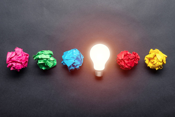 Lightbulb and crumpled colorful paper balls on black background. Successful solution of problem. Idea generation and brainstorming. Genius idea among failing ideas metaphor. Business motivation - Photo, image