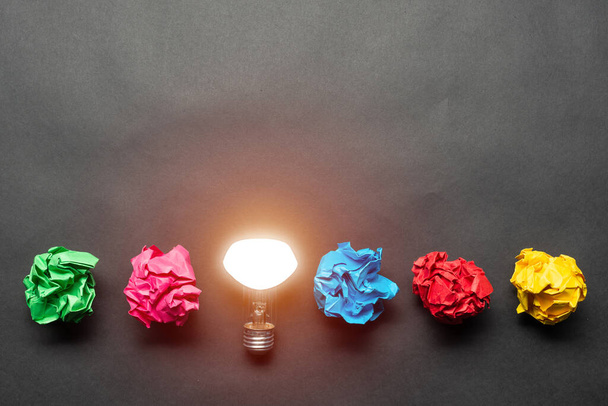 Lightbulb and crumpled colorful paper balls on black background. Successful solution of problem. Idea generation and brainstorming. Genius idea among failing ideas metaphor. Business motivation - Photo, image
