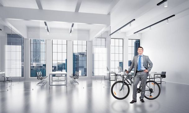 Smiling man in business suit standing with bike at comfortable conference room. Happy businessman with bicycle at modern office interior with big windows. Loft coworking space advertising. Mixed media - Zdjęcie, obraz