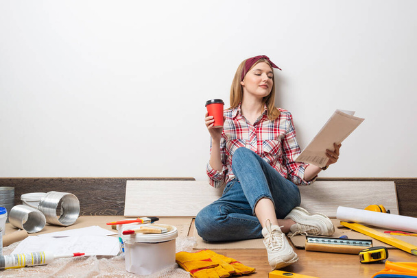Smiling girl sitting on floor and holding paper blueprint. Home remodeling and house interior redesign. Construction tools and materials for building. Young attractive woman wearing casual clothes - Photo, image
