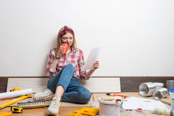 Happy pretty girl sitting on floor with paper blueprint. Home remodeling and house interior redesign. Construction tools and materials for building. Young woman studying renovation project of flat. - Foto, immagini