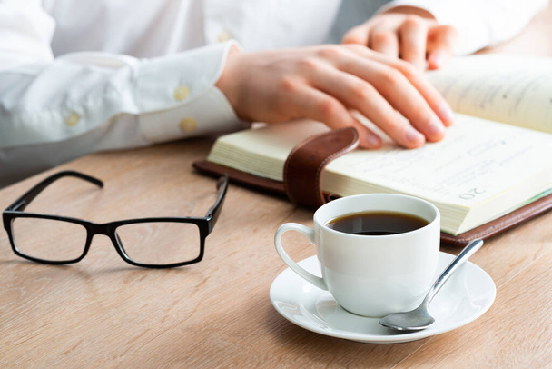 Mężczyzna w białej koszuli czyta notatki w notatniku na drewnianym stole. Biznesmen w miejscu pracy z filiżanką kawy. Planowanie biznesowe i organizacja dnia pracy. Zbliżenie męskich dłoni z przypomnieniem. - Zdjęcie, obraz
