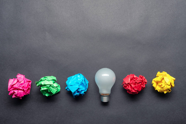 Lightbulb and crumpled colorful paper balls on black background. Successful solution of problem. Idea generation and brainstorming. Genius idea among failing ideas metaphor. Business motivation - 写真・画像