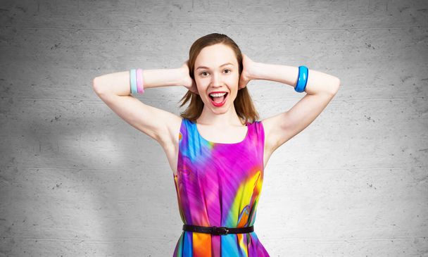 Redhead beautiful girl closed her ears with hands. Joyful lady with charming smile in bright colorful dress and bracelets against grey wall background. Portrait of happy young woman smiling broadly - Foto, Bild
