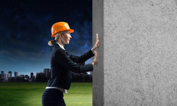 Beautiful woman architect pushing concrete wall. Young specialist in safety helmet on night city background. Sale building material and services. Grey concrete wall as banner with copy space. - Фото, зображення