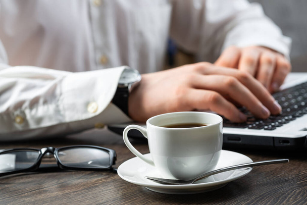 Businessman sitting at desk and using laptop. Close-up of man hands typing on keyboard in office. Side view consultation workplace with glasses and coffee cup. Business experience and professionalism. - Photo, Image