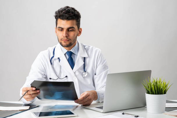 Handsome young doctor looking at x-ray scan. Physician in white coat with stethoscope sitting at desk with laptop. Professional medical diagnosis and treatment in hospital. Medical healthcare services - Φωτογραφία, εικόνα