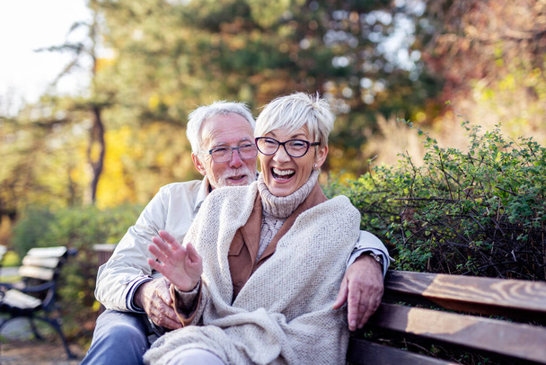 Mature couple sitting on bench in public park talk and smile - Fotó, kép