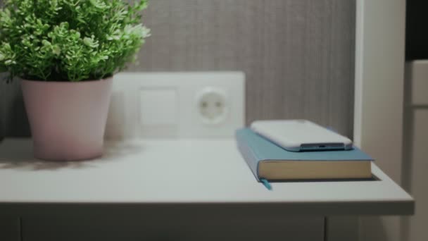 Vrouwelijke hand uit te schakelen een wekker op mobiele telefoon op nachtkastje met indoor plant en boek - Video