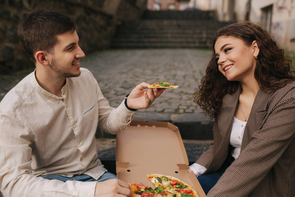 Smiling love couple eating vegan pizza at street on stairs. Handsome man give slice of pizza to woman - Fotó, kép