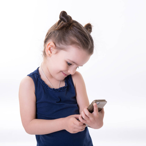 Portrait of cool cheerful little girl taking photo with smart phone. Cute kid shooting selfie on front camera isolated on white background. Child using a mobile phone. - Fotó, kép