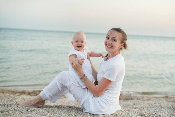 Young beautiful mother and baby sitting on sandy beach looking at camera and smile. - Photo, Image