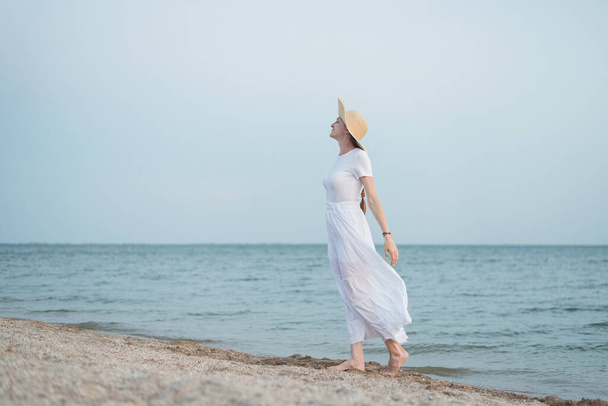Gyönyörű fiatal nő fehér ruhában és kalapban a parton. Arisztokrata lány a tengerparton. - Fotó, kép