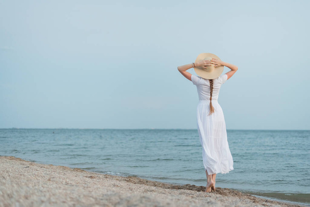 Delicate young woman in white dress and hat walks on sea shore. Back view. - Foto, afbeelding