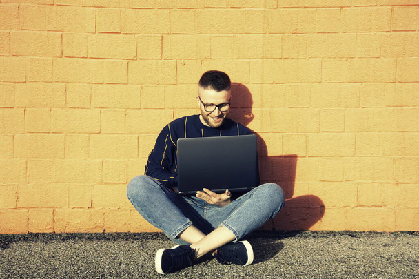 Fiatal kaukázusi férfi alkalmi viselet ül keresztlábú a földön, és dolgozik a laptop - Freelancer dolgozik kívül gépelés laptop számítógép, Intelligens munka koncepció - Vörös fal a háttérben - Fotó, kép