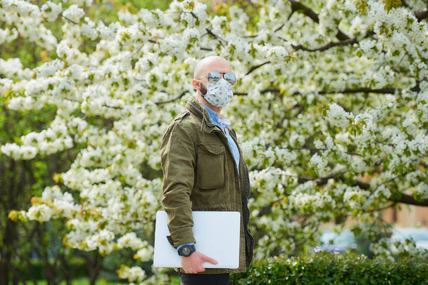 A bald man with a beard in a medical face mask to avoid the spread coronavirus walks with a laptop in the park. A guy wears n95 face mask and a pilot sunglasses in the garden between flowering trees. - Photo, Image
