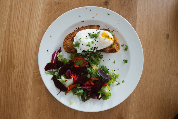 Tojásos buggyantott tojás egy darab kenyéren zöld salátával egy tányéron és a fa asztalon. Egészséges reggeli - Fotó, kép