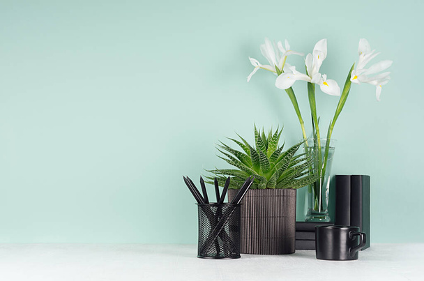 Elegant workplace with black stationery, books, coffee cup, green houseplant of aloe, fresh white iris bouquet on green mint menthe wall and white wood table. - Φωτογραφία, εικόνα