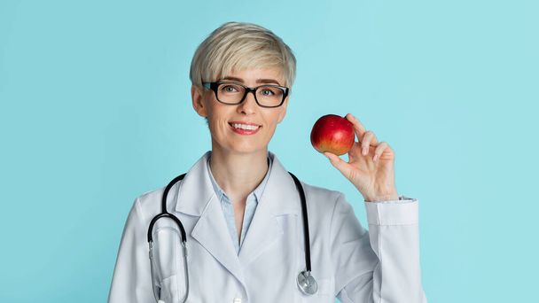 Healthy eating concept. Adult woman doctor in glasses holds red apple in hand - Photo, Image