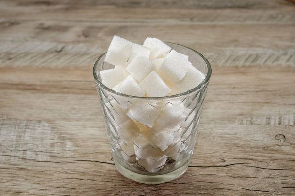 A glass of sugar crystals or cubes on a wooden table. Intake of bad calories. - Foto, Imagen
