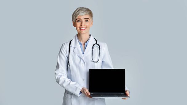Mosolygó női orvos fehér kabátban azt mutatja, egy laptop egy üres képernyőn. - Fotó, kép