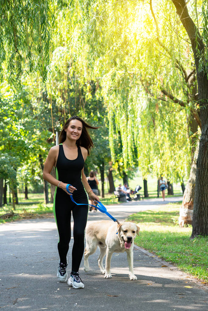 Cheerful pretty young woman walking and running with her dog in summer park. Active leisure, beautiful brunette girl at morning  jogging with puppy - 写真・画像