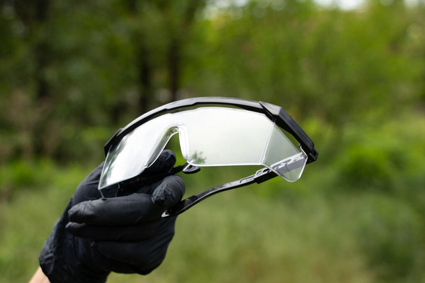 χέρι σε μαύρο ιατρικά γάντια κρατά ιατρικά γυαλιά σε ένα απομονωμένο φόντο - Φωτογραφία, εικόνα