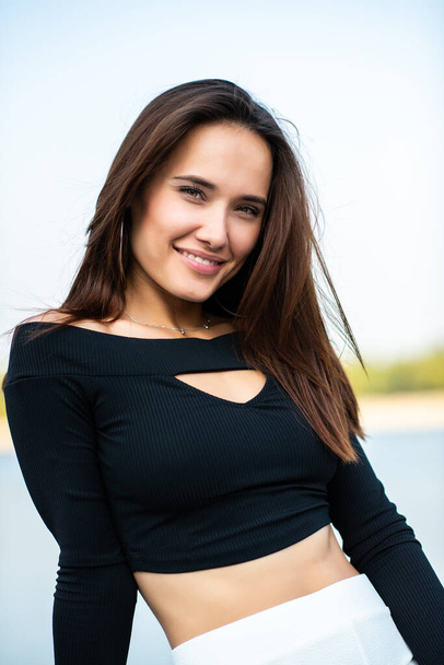 Charming brunette girl smiling outdoor. Young woman, summer portrait. Female model posing over nature background with river, beach and blue clean sky. - Фото, зображення