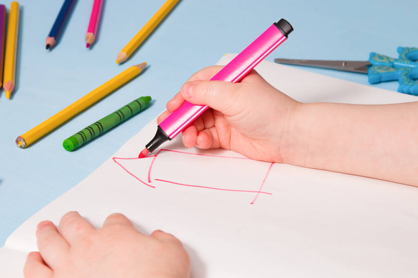 a small child painstakingly paints with a pink felt-tip pen a house in an album, a copy place, a top view, a blue background, what to do with a quarantined child, activities for children - Фото, изображение