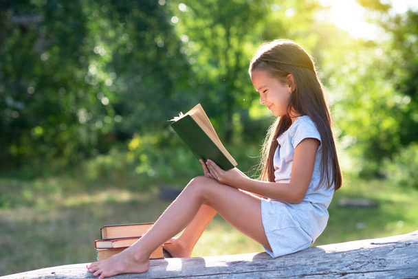 Charming little girl with long brown hair reads book outdoor sitting on tree in summer park or in a forest glade. Child booklover - Foto, Imagem